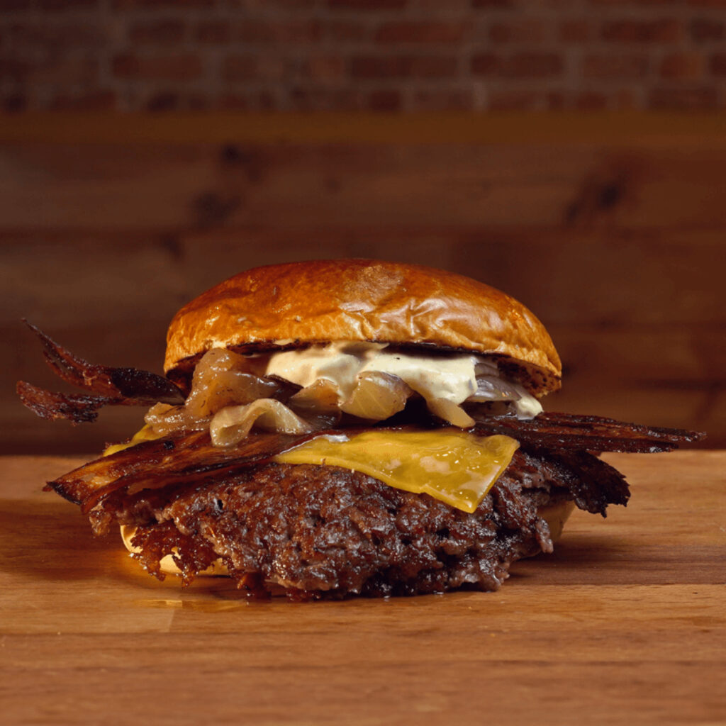 Hamburguesa clásica estilo americano, viene con 2 carnes, smashed, queso cheddar, cebolla salteada al momento, bacon y mayonesa y pepinillo en Madrid